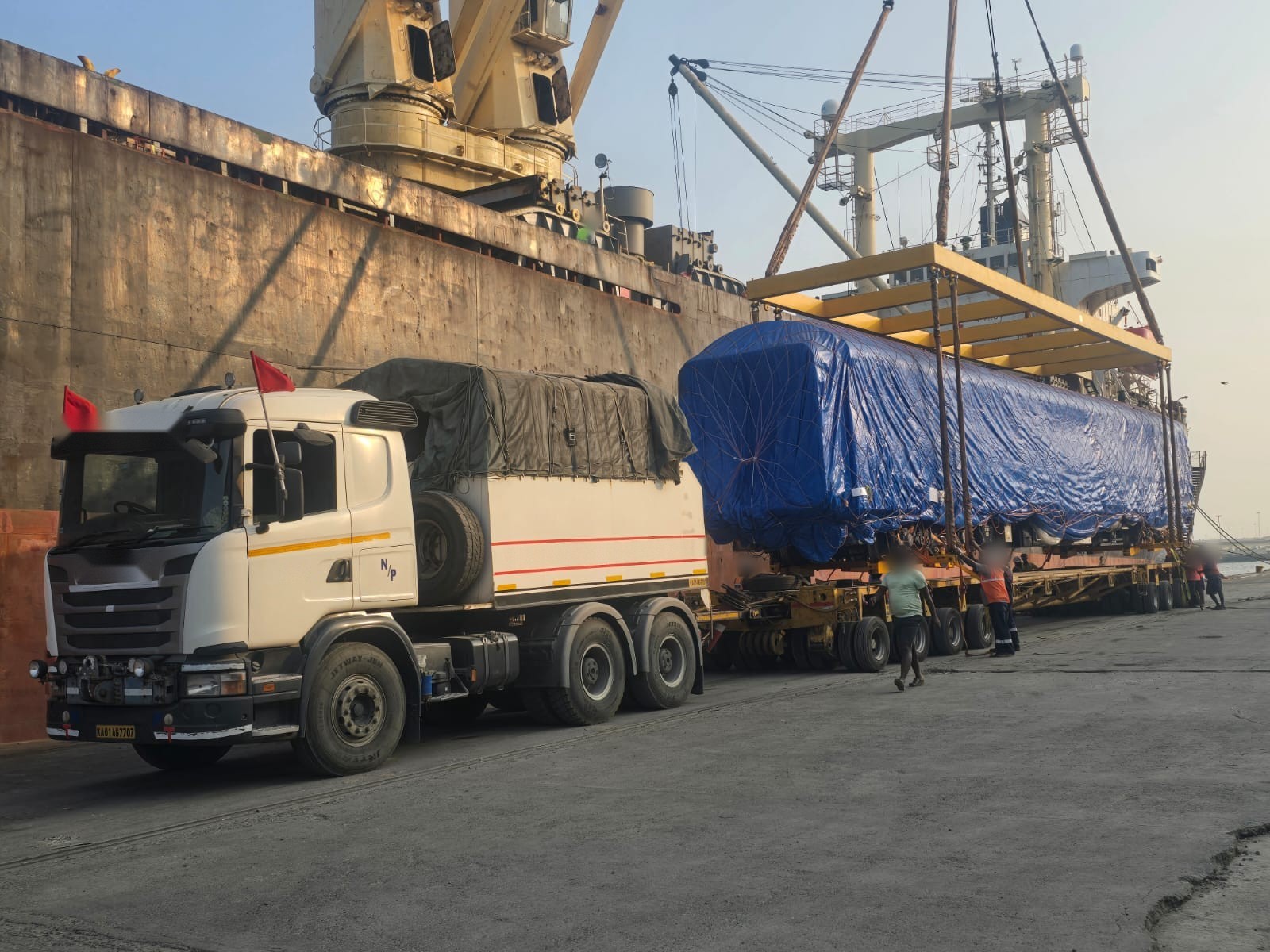 Tarpaulin-covered long cargo on hydraulic modular axles alongside a geared vessel, ship crane rigged overhead and crew guiding the set-down.